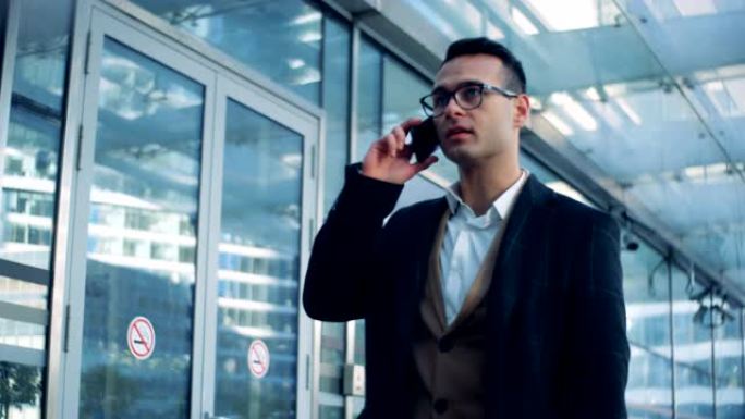 男性专家正沿着办公楼的玻璃墙行走，并在电话中交谈。6k电影摄像机镜头。