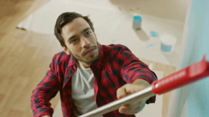 一个穿着棕色牛仔裤和红色格子衬衫的男人的特写镜头，用滚轴画了一堵墙。油漆颜色为浅蓝色。家里的房间装修