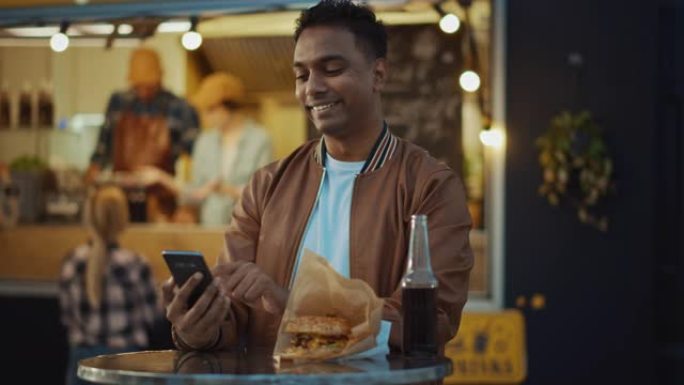 英俊的年轻印度男子坐在户外街头美食咖啡馆的桌子旁，正在使用智能手机。他正在浏览互联网或社交媒体，发布