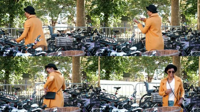 年轻的黑人妇女穿着黄色的未扣黄色豌豆外套，黑色的帽子和墨镜停放她的自行车，穿上斜挎包，然后走开，没有