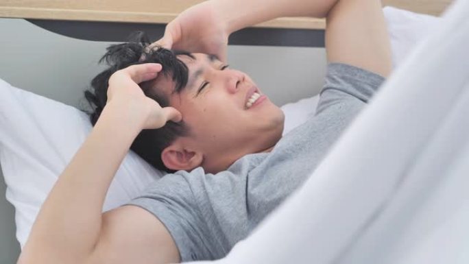 亚洲男人感到压力，双手抱头，感到头痛，躺在床上。生病在家