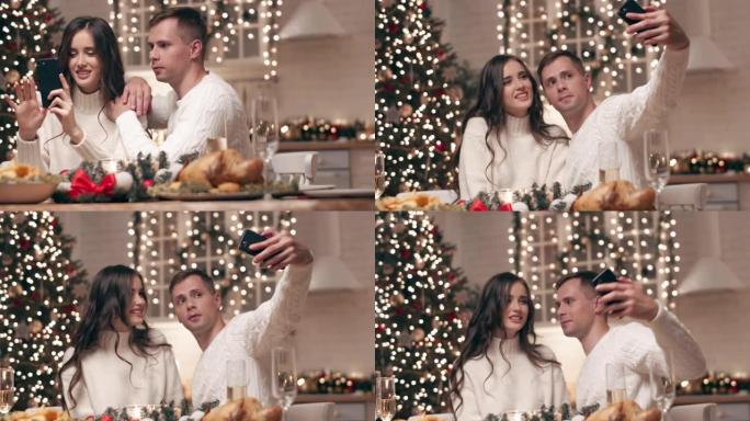 美丽的年轻夫妇坐在圣诞餐桌旁等待朋友。年轻人在手机摄像头上联合自拍，他们将圣诞节照片发送给亲朋好友。