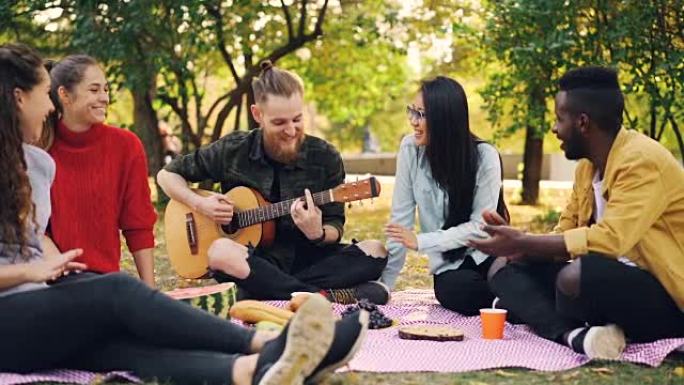 快乐的学生在公园里弹吉他和享受音乐的慢动作在秋天的野餐中，吉他手正在演奏，而他的朋友们正在拍手。