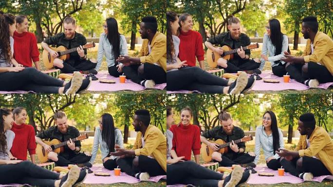快乐的学生在公园里弹吉他和享受音乐的慢动作在秋天的野餐中，吉他手正在演奏，而他的朋友们正在拍手。
