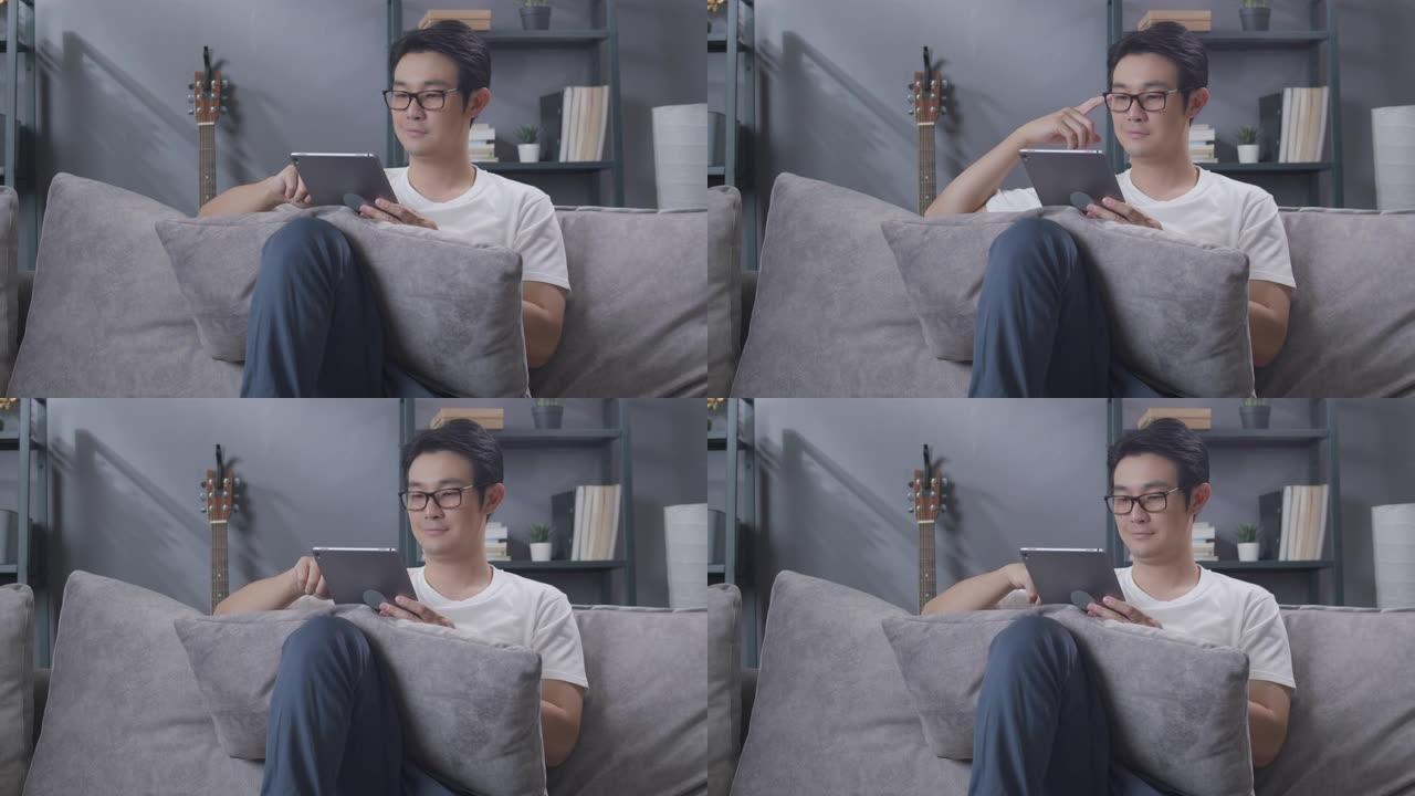 亚洲人坐在家里的客厅沙发上放松时，在数字平板电脑上阅读新闻。戴眼镜的成年男子在网上看报纸，在他的房子