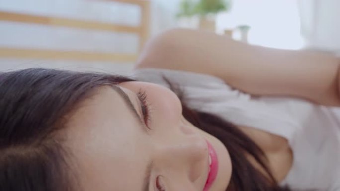 年轻的亚洲妇女使用智能手机检查社交媒体感到快乐的微笑，躺在床上后早上醒来，美丽迷人的日本女孩微笑在家