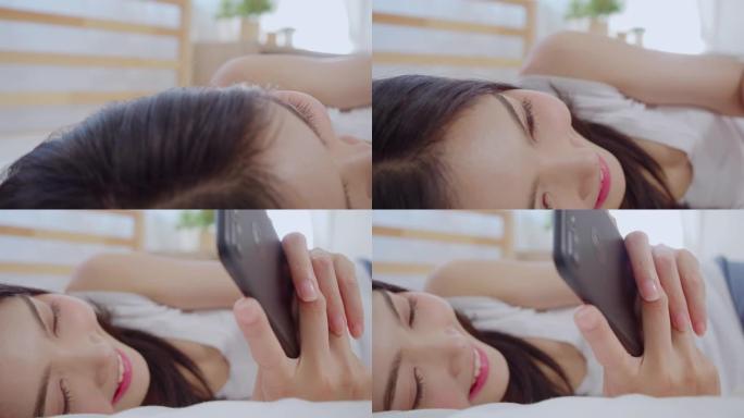年轻的亚洲妇女使用智能手机检查社交媒体感到快乐的微笑，躺在床上后早上醒来，美丽迷人的日本女孩微笑在家