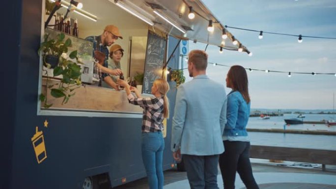 Food Truck员工向快乐的时髦顾客提供牛肉汉堡，薯条和冷饮。人们在外面的餐桌上吃饭。商用卡车在