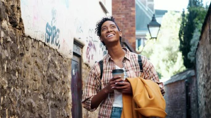 穿着格子衬衫的年轻黑人妇女站在小巷里，拿着外套和外卖咖啡，对着镜头微笑，特写镜头