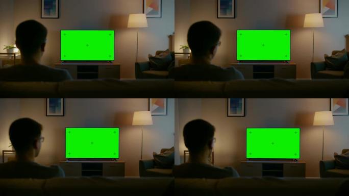 戴眼镜的年轻人坐在沙发上，看着电视，上面放着水平的绿色屏幕模型。现在是晚上，家里的房间有工作灯。