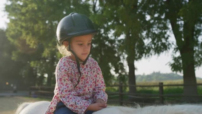 真实的近距离拍摄一个可爱的小女孩与骑师头盔是骑着一匹白马在骑马厩与阳光