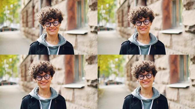 美丽女孩的慢动作肖像是眼镜微笑着看着摄像机站在户外的大城市的街道上。人、情感和城镇概念。