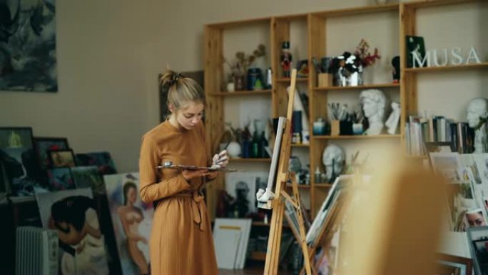 漂亮女孩画家独自在工作室里工作，拿着调色板和画笔，看着画布。职业、创意青年和工作空间概念。