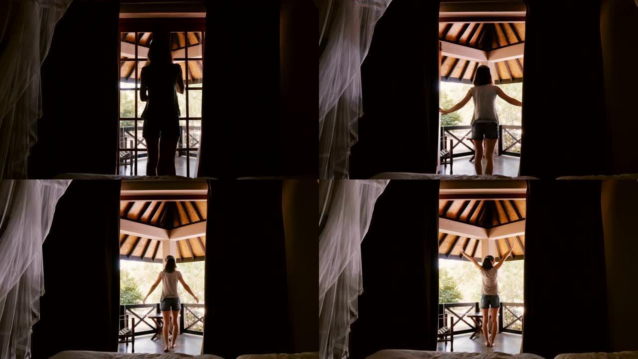 后视图快乐的年轻旅游女人打开小屋窗帘和窗户门，走出去在阳光明媚的露台伸展。