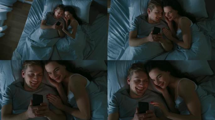 幸福的年轻夫妇晚上在床上使用智能手机，他们共享屏幕，浏览社交网络，共享图片，使用互联网并获得乐趣。俯