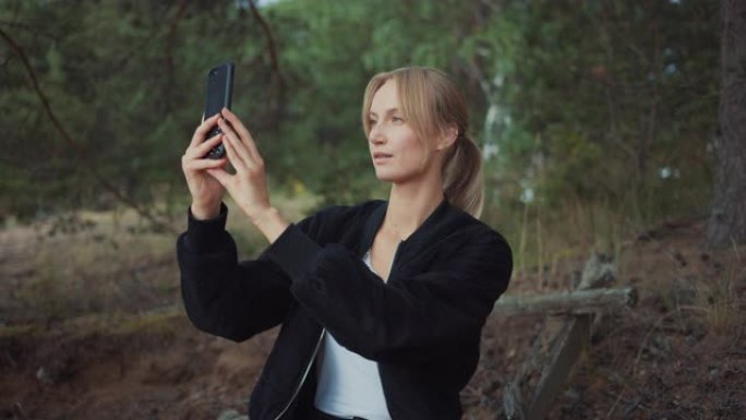 一个年轻美丽的金发女人在浪漫的自然氛围中的肖像。女孩穿着黑色衣服，正在用手机拍照或自拍。她坐在松树林