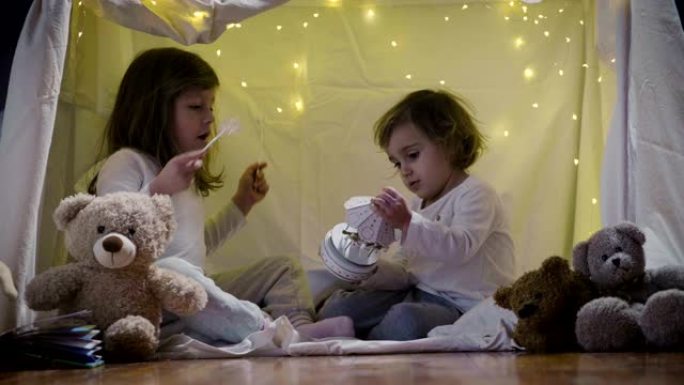 两个有妈妈的小女孩晚上在家里玩，搭个野营帐篷，用手电筒看书，在里面睡觉。