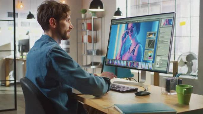 穿着牛仔裤衬衫的男性数字编辑器在他的个人电脑上大显示屏上的照片编辑软件中工作。他在凉爽的办公室阁楼工