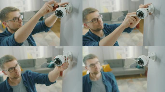 戴着眼镜的年轻人穿着蓝色衬衫，正在家里的白色墙壁上拧一个带有两个天线的现代wi-fi监控摄像头。