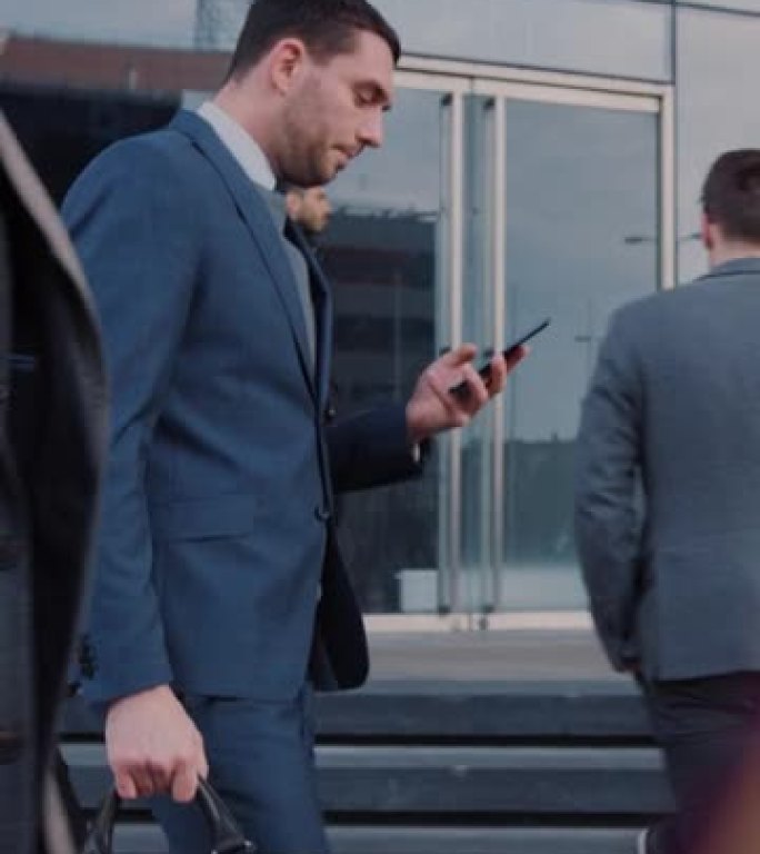 穿着西装的高加索商人晚上在黑暗的街道上使用智能手机。其他办公室人员走过。垂直屏幕方向9:16的视频素