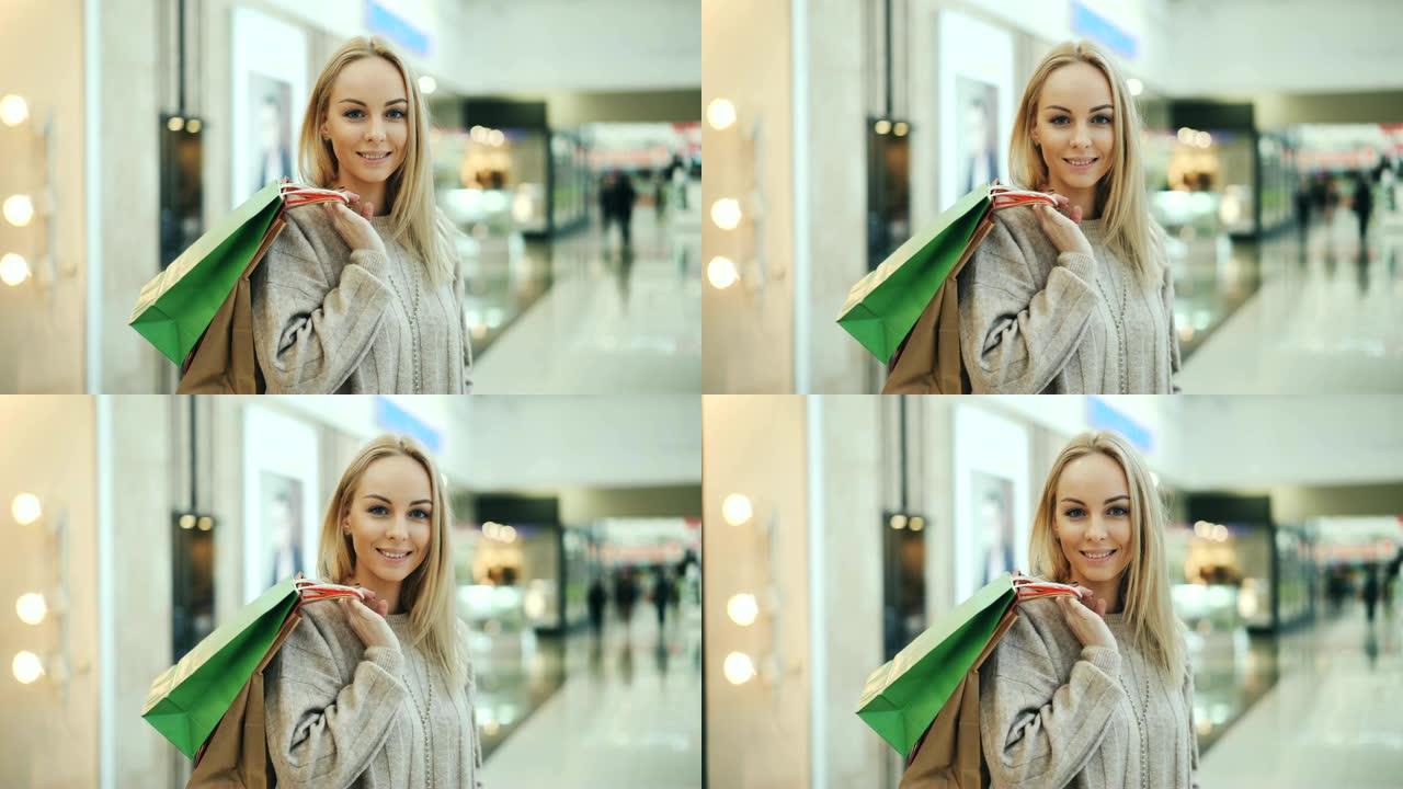 长着金色长发的漂亮女士的肖像看着相机，微笑着站在纸袋的购物中心里。年轻人和买东西的概念。