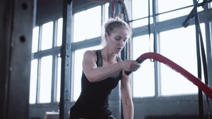 年轻美丽的运动金发白人妇女用战斗绳锻炼，在大型健身房慢动作中休息。