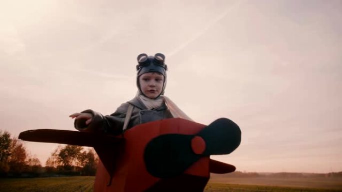 快乐的小男孩穿着有趣的纸板飞机服装在日落秋野外旋转，玩航空飞行员慢动作。