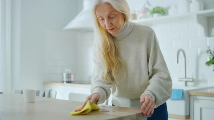 有爱心的高级家庭主妇擦拭木制厨房桌子上溢出的咖啡或茶。美丽的老年女性使用家用清洁剂擦拭。养老金领取者