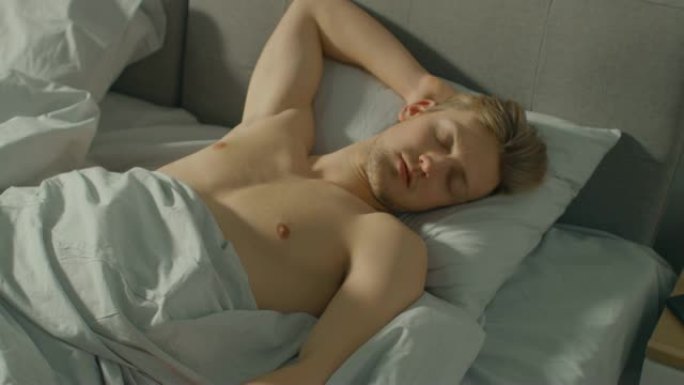英俊的金发男孩舒适地睡在床上。年轻的白种人正在熟睡。清晨的阳光透过窗户