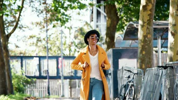 时尚的年轻黑人妇女戴着帽子，太阳镜，蓝色牛仔裤和黄色豌豆大衣，沿着树木环绕的街道走向相机，戴着耳机听