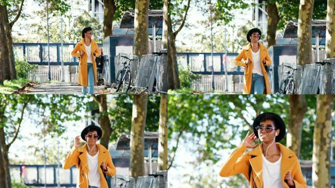 时尚的年轻黑人妇女戴着帽子，太阳镜，蓝色牛仔裤和黄色豌豆大衣，沿着树木环绕的街道走向相机，戴着耳机听