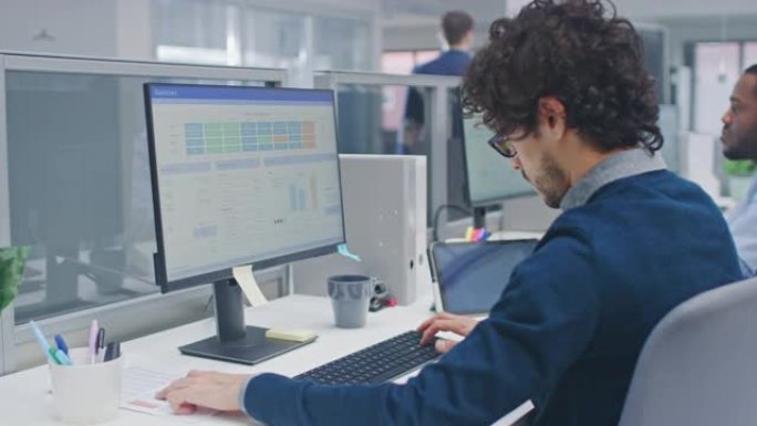卷发的年轻英俊经理在台式计算机上工作，并将数据与软件进行比较。在现代开放式办公室中，多元化且积极进取