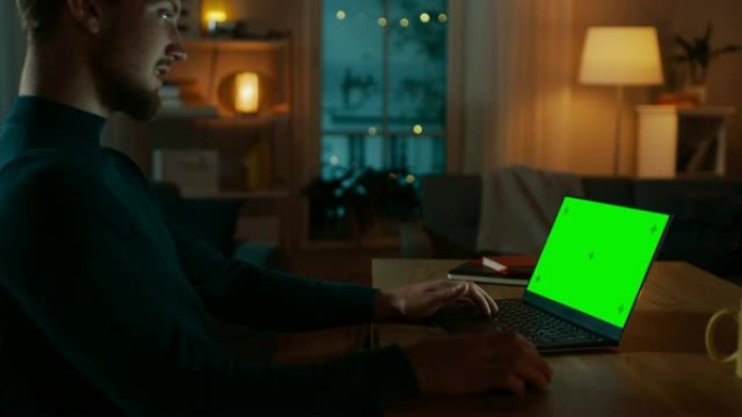 坐在办公桌前的男人在带有绿色模拟屏幕的笔记本电脑上工作。深夜在他的客厅里，男人使用笔记本电脑。侧视放