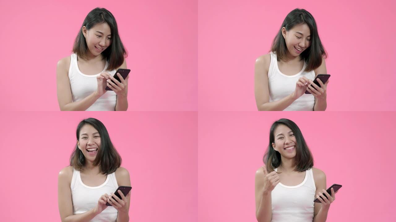 使用智能手机检查社交媒体的年轻亚洲女性在粉红色背景下穿着休闲服装时感到快乐的微笑。快乐微笑可爱的快乐