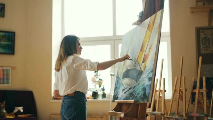 漂亮女孩画家在调色板上混合颜料，然后在画布上绘画海景，创造美丽的图画。漂亮的现代工作室与工具是可见的