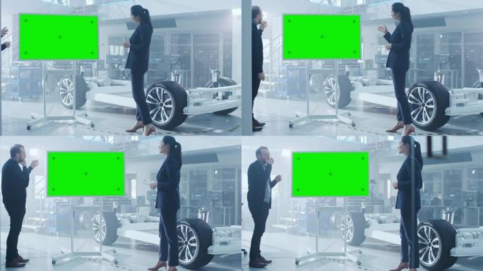 男性和女性设计工程师正在电动汽车底盘原型旁边的带有绿色屏幕的交互式白板上讨论一些事情。在带有车架的高