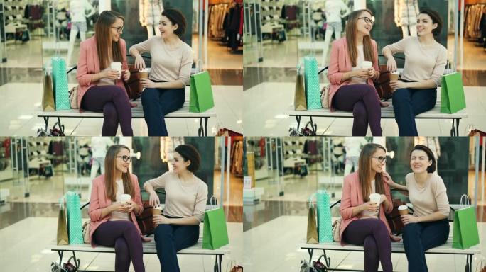 快乐的女孩正在社交，坐在购物中心的长凳上聊天和笑，那里有去的饮料和购物袋。可见有服装的商店橱窗。