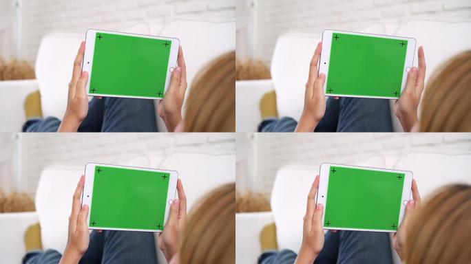 年轻的亚洲妇女使用绿色屏幕的黑色平板电脑设备。亚洲妇女拿着平板电脑，坐在客厅的沙发上滚动页面。色度键