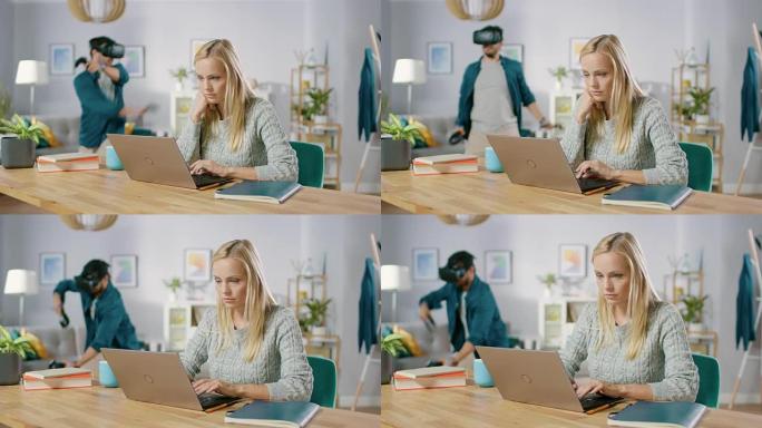美丽集中的女人在她的桌子上的笔记本电脑上工作，在她的男朋友后面戴着虚拟现实耳机玩带有控制器的视频游戏