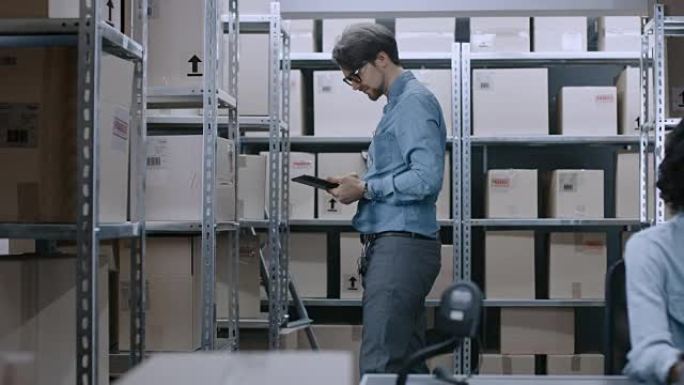 仓库工人使用数字平板电脑检查货架上的库存，以获取正确的纸板箱包装。在后台，他的女同事在个人电脑上工作