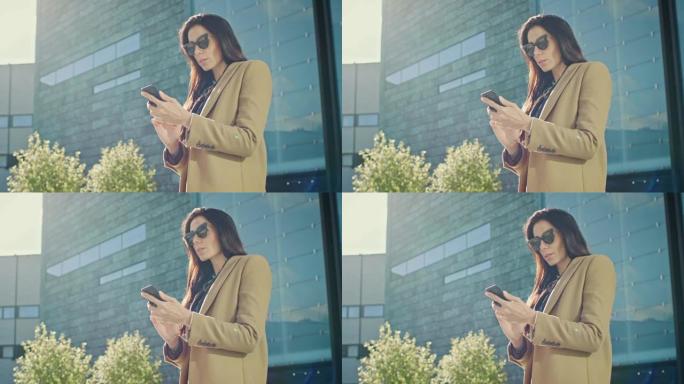 优雅的女商人站在现代玻璃建筑附近时，使用智能手机开展业务。穿着外套和墨镜的美丽时尚女性在现代城市环境