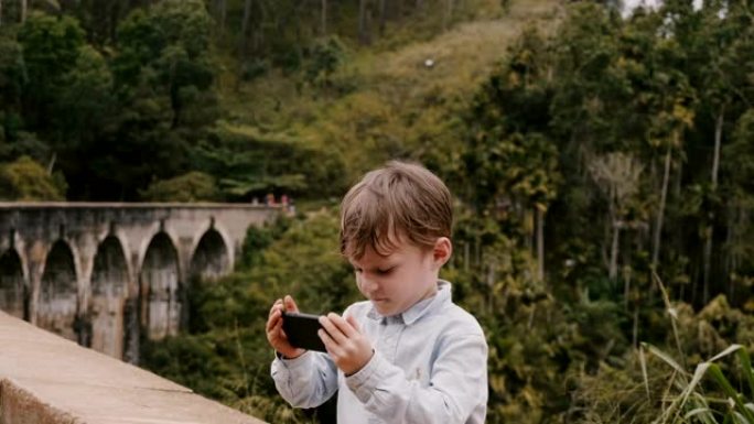 斯里兰卡度假时，快乐可爱的欧洲小男孩在埃拉九拱桥拍摄智能手机照片。