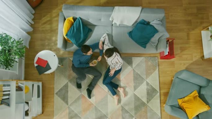 幸福的夫妇坐在地板上聊天。女孩给一个年轻人带了一杯咖啡。舒适的客厅，配有现代室内地毯、沙发、椅子、桌