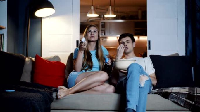 快乐的年轻新婚白人家庭坐在家里的沙发上看电影，零食在慢动作。