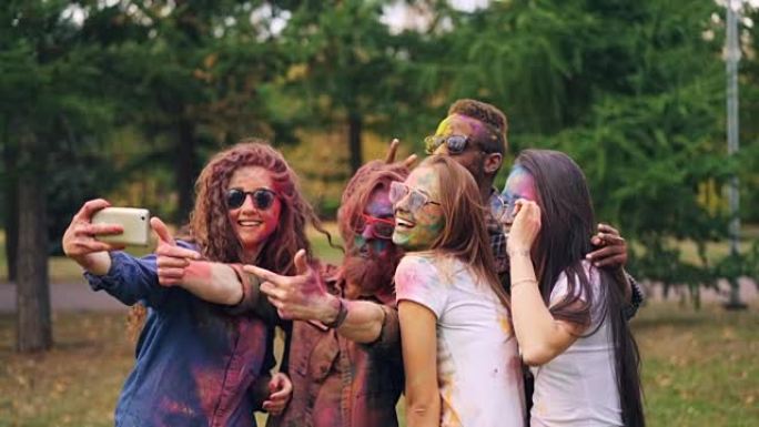 脸上和头发彩绘的快乐男女朋友在洒红节上使用智能手机摄像头自拍，人们摆姿势并微笑着。乐趣和技术概念。