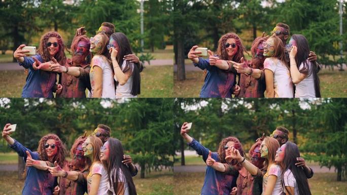 脸上和头发彩绘的快乐男女朋友在洒红节上使用智能手机摄像头自拍，人们摆姿势并微笑着。乐趣和技术概念。