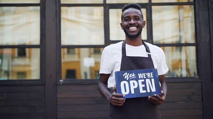快乐的非洲裔美国商人咖啡馆老板的慢动作肖像拿着我们是站在外面看着相机的开放标志。小企业和青年概念。