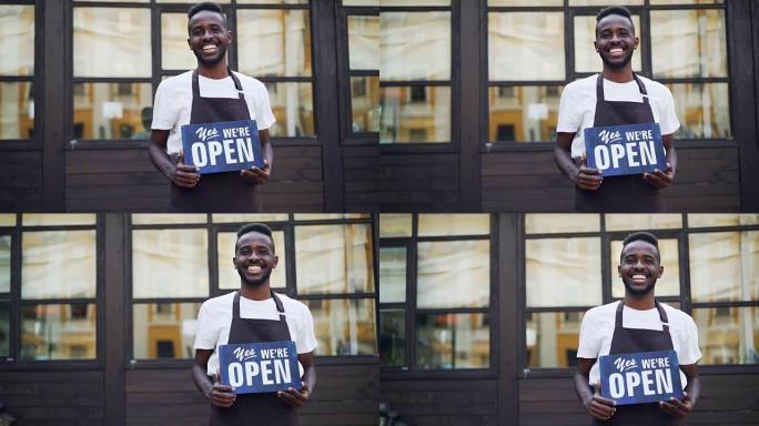 快乐的非洲裔美国商人咖啡馆老板的慢动作肖像拿着我们是站在外面看着相机的开放标志。小企业和青年概念。