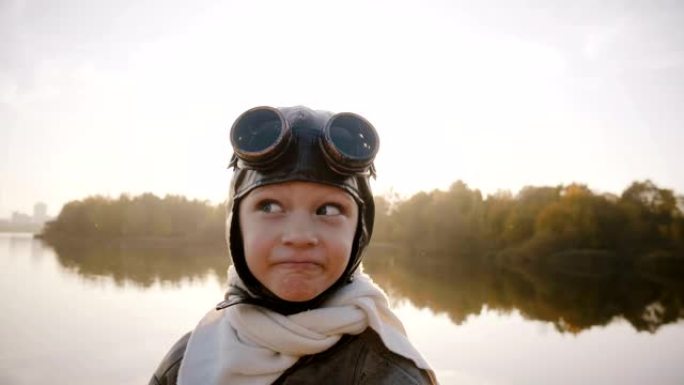 穿着老式飞行员服装的小男孩，戴着围巾和眼镜，看着相机，在湖慢动作附近制作有趣的面孔。