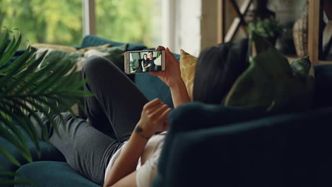 社交女孩的后视图使用智能手机进行在线视频通话躺在舒适的房子的沙发上，人们在说话和打手势。对话和互联网